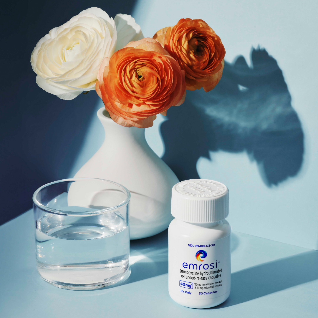 Emrosi bottle beside flowers, a water glass, and a cosmetic jar
